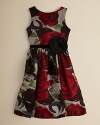 A fun party dress in a bold red floral, accented with a delicate velveteen sash and large flower at the hip.